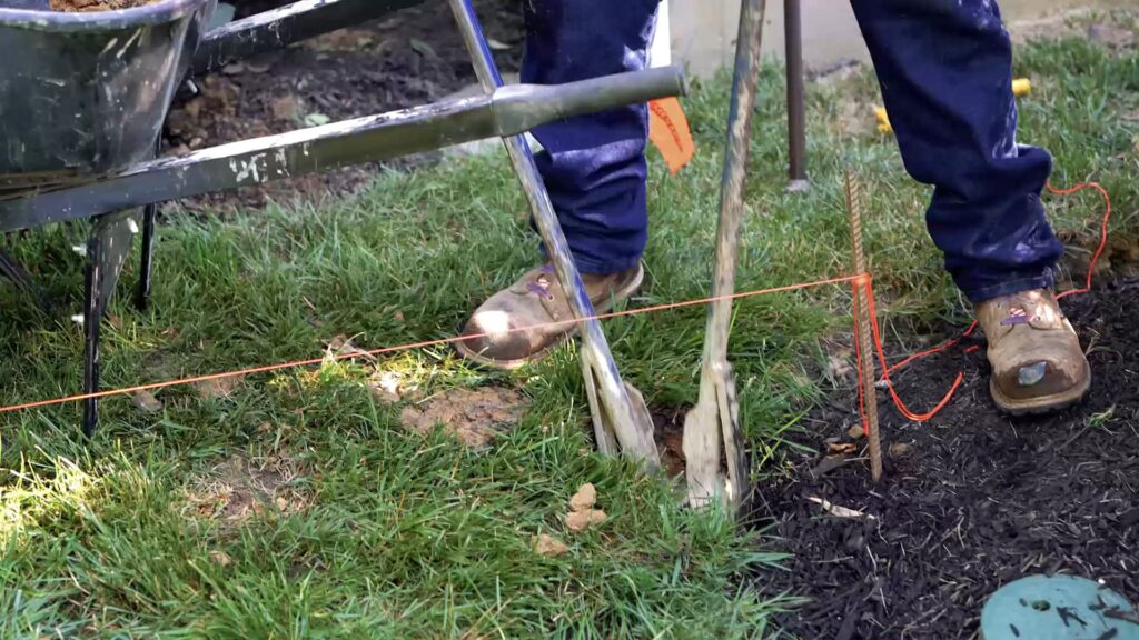 Still frame from a video recording of a man in construction clothes using a post shovel in a grassy yard next to a wheelbarrow and mulch bed